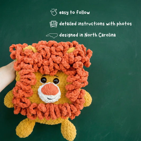 Hand holding a plush crochet lion amigurumi with a fluffy orange mane against a green background.