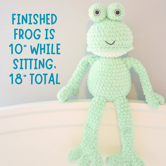Plush crochet frog toy with large eyes, sitting on a white surface, amigurumi pattern display.