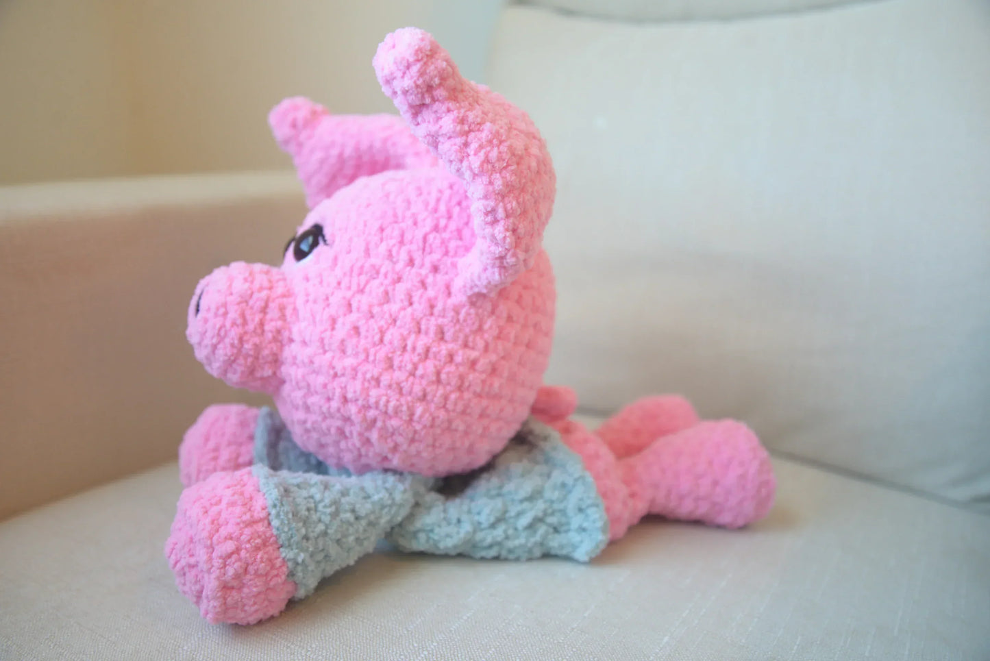 Pink crochet pig snuggler plush toy with blue blanket on a cream chair