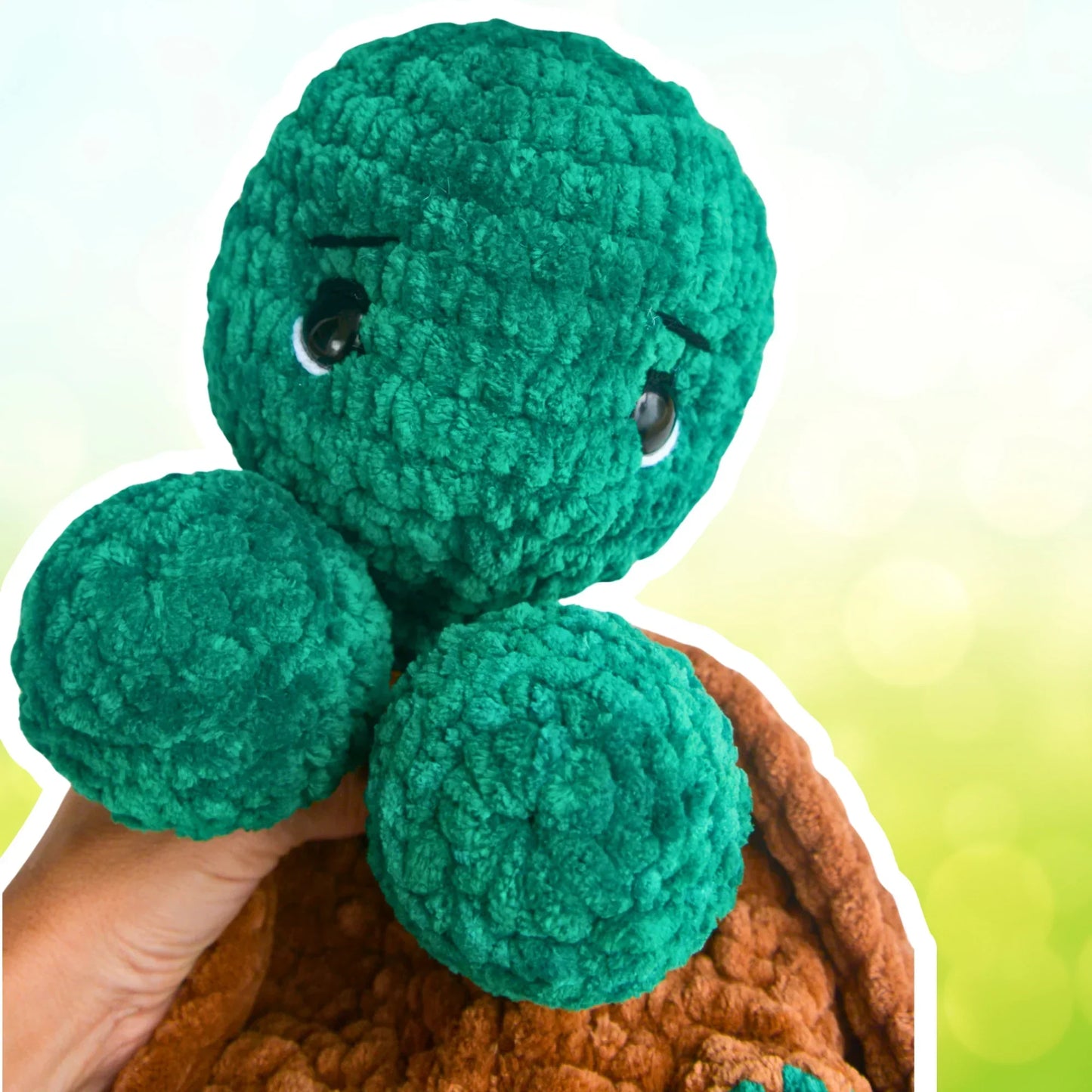 green crochet turtle plushie with brown shell, held by hand against soft pastel background