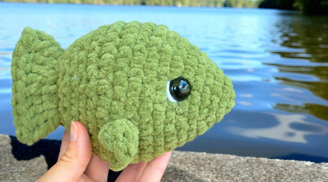 Hand holding a green crochet fish plushie near a lake, beginner amigurumi stuffed animal pattern.