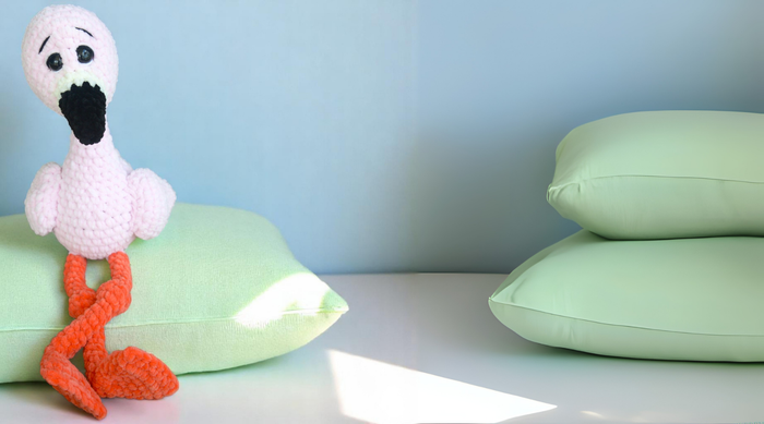 Crochet flamingo plushie with orange legs sitting on green pillow, pastel background