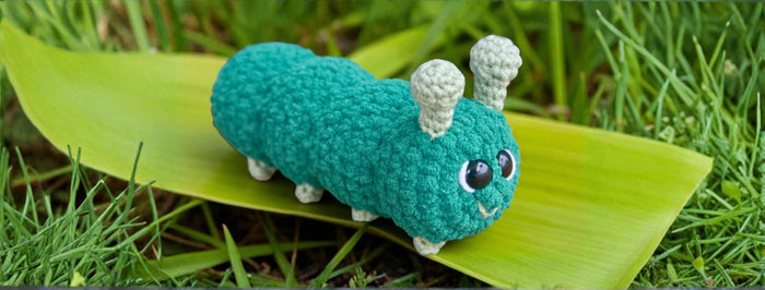 Cute crocheted teal caterpillar toy with white antennae and big eyes on green leaf