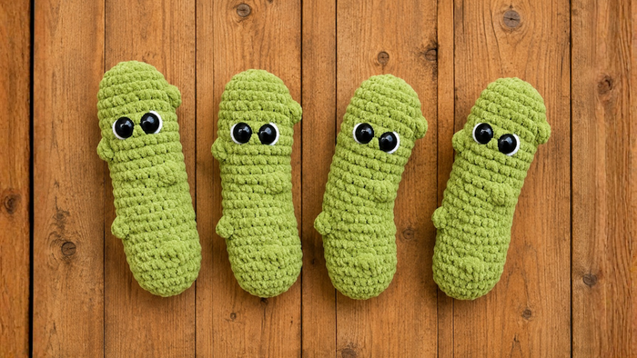 Four handmade green crochet plush toys resembling pickles with large eyes on a wooden background