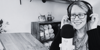 Woman with glasses recording a crochet podcast, yarn and crochet hooks in background