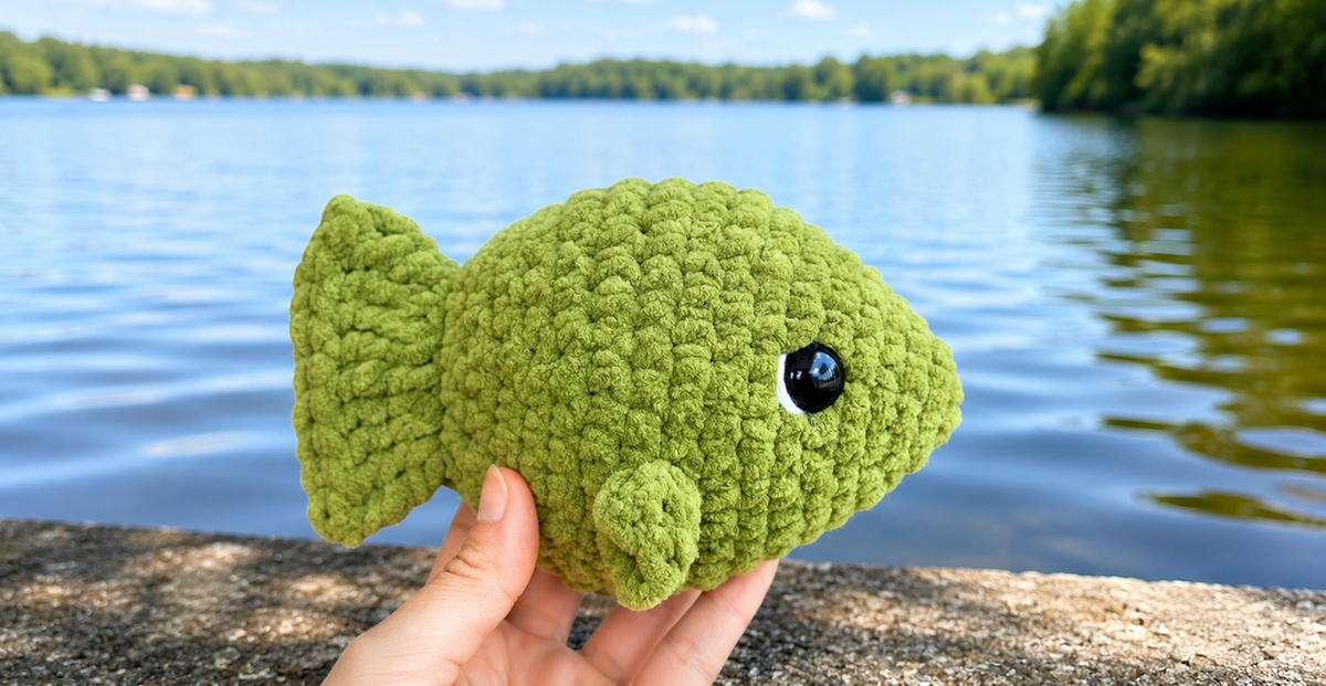 Hand holding a green crochet fish plushie by a lake, amigurumi stuffed animal toy