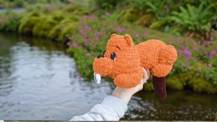 Hand holding orange crochet beaver plushie by a river with green plants and purple flowers.