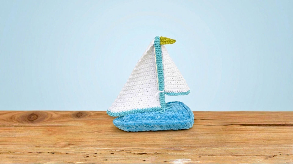 crochet sailboat amigurumi with white sail and blue base on wooden table, light blue background