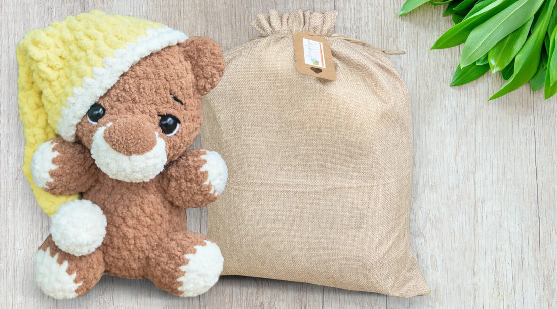 Handmade crochet teddy bear with yellow hat next to burlap sack on wooden surface with green leaves