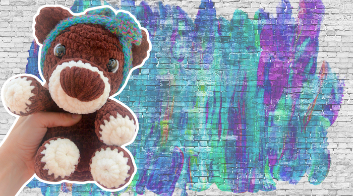 Hand holding a brown and white crocheted teddy bear with a blue-green headband against a colorful graffiti brick wall