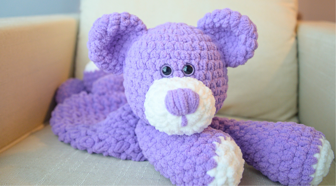 Handmade purple crochet teddy bear with white snout and paws, lying on a beige sofa