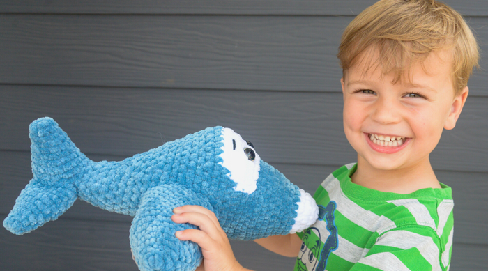 Smiling boy in green striped shirt holding a handmade blue crochet shark toy against gray wall