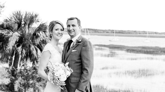 Smiling bride and groom in wedding attire posing outdoors by palm trees and water