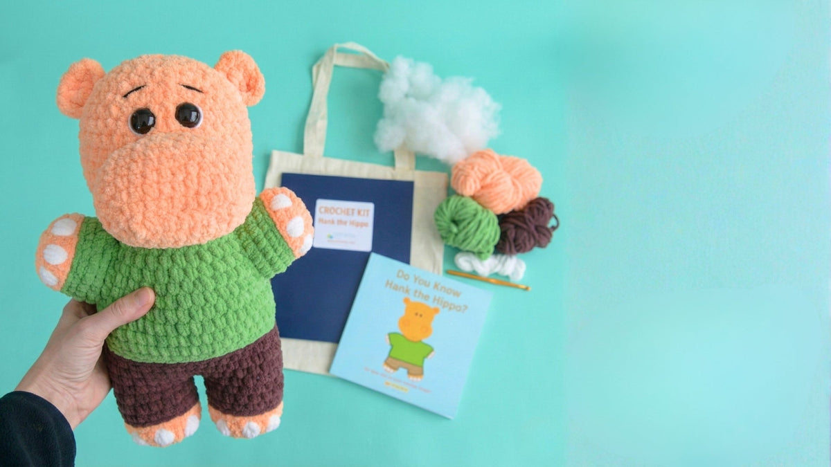 Beginner crochet kit with plush hippo toy, yarn, crochet hook, book, and tote bag on teal background