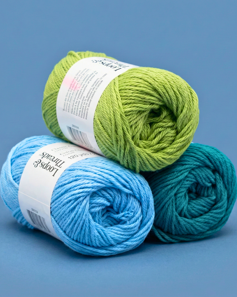 Stack of green, blue, and teal crochet yarn skeins for amigurumi projects on blue background