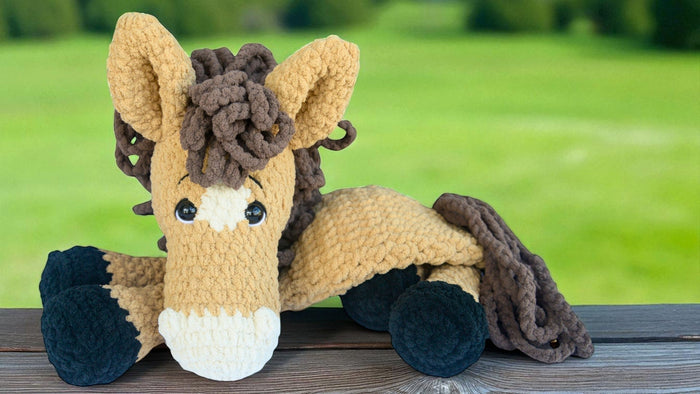 Crochet horse plushie with big eyes and textured mane on wooden surface, green field background