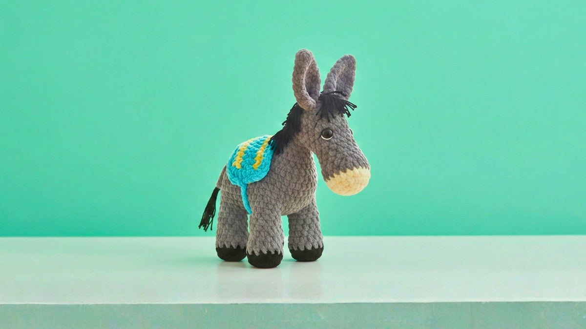 Hand holding gray crocheted amigurumi donkey with blue saddle on light blue background