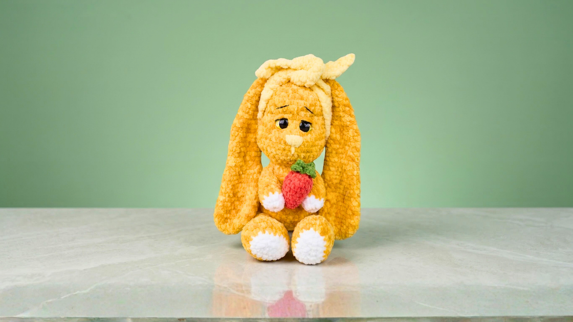 Yellow crochet bunny plushie holding a strawberry, amigurumi toy on marble surface