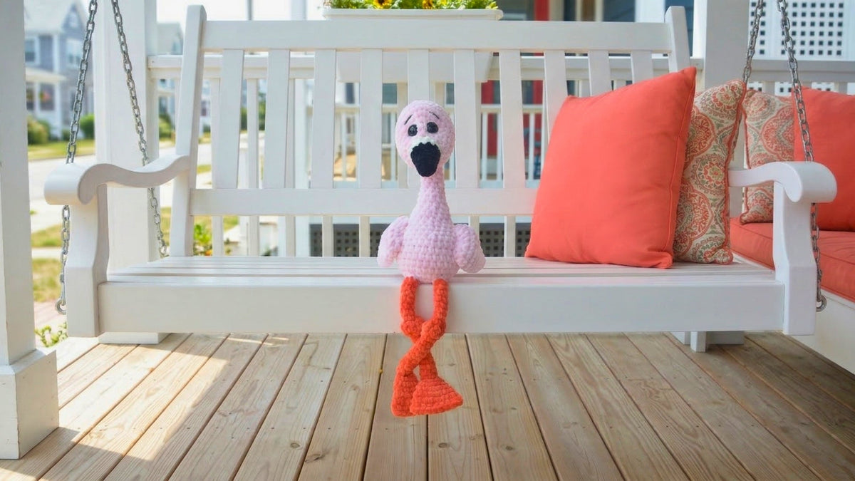 Pink crochet flamingo plushie on white porch swing with coral pillows, amigurumi toy pattern