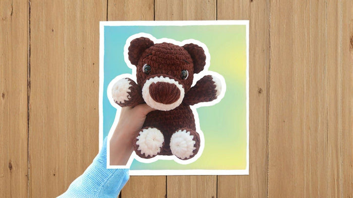 Hand holding a crochet teddy bear plushie, brown with white accents, against blue background.