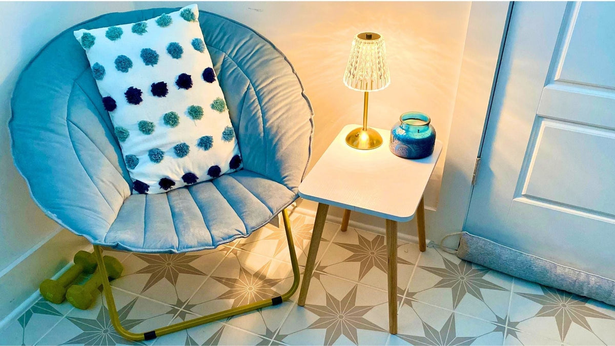 Cozy corner with a blue papasan chair, pom pom pillow, side table, lamp, and blue candle.