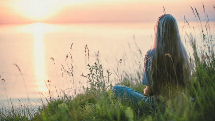 Woman with long hair sitting on grass by lake, watching sunset with soft pink sky