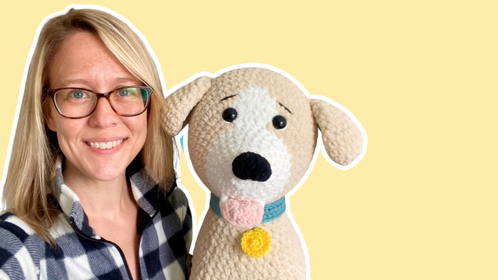 Smiling woman wearing glasses and plaid jacket with handmade crochet dog toy on yellow background