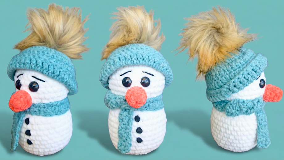 Three crocheted snowmen with blue hats, scarves, orange noses, and faux fur pom-poms on wooden surface with snow