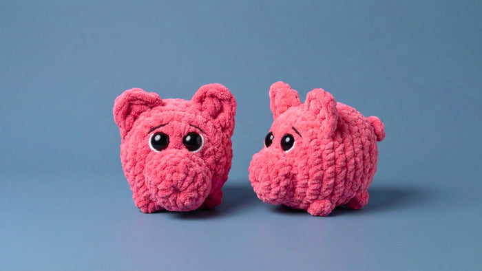 Two pink crocheted plush pigs with big shiny eyes held in hands against blurred colorful background