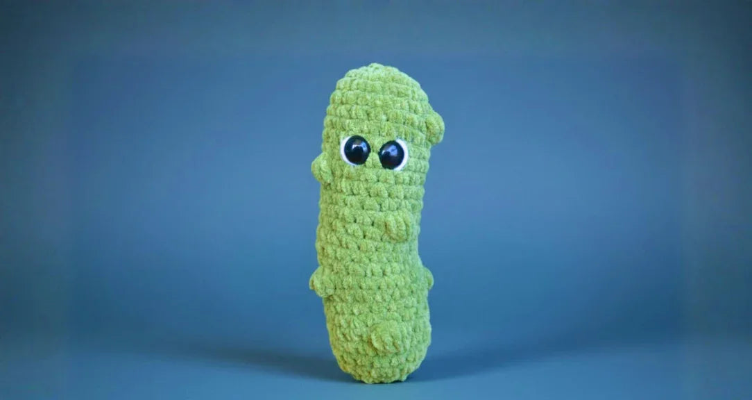 Four handmade green crochet plush toys resembling pickles with large eyes on a wooden background