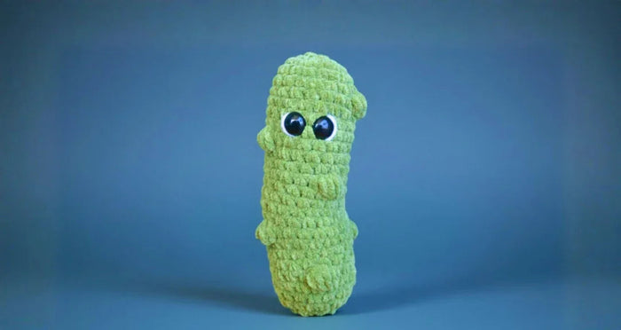 Four handmade green crochet plush toys resembling pickles with large eyes on a wooden background