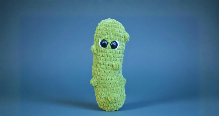 Green crochet pickle plushie with big safety eyes, soft texture, amigurumi toy on blue background