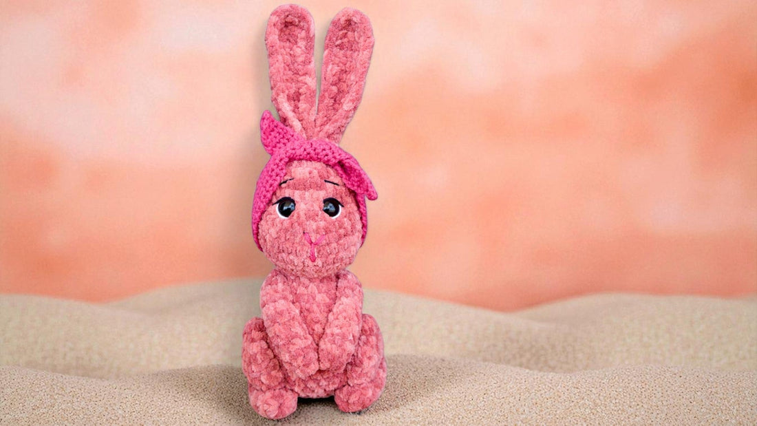 Pink plush bunny toy with knitted headband sitting on beige textured surface against peach background