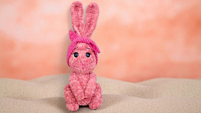 Pink plush bunny toy with knitted headband sitting on beige textured surface against peach background