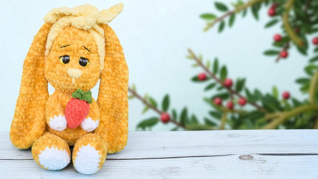 Handmade orange crochet bunny holding a red carrot on white wooden surface with blurred green leaves background