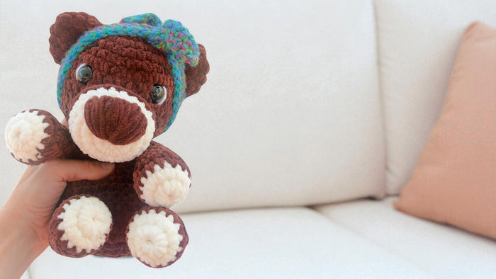 Hand holding a brown and white crocheted teddy bear with a colorful headband on a white couch