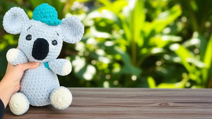 Hand holding a handmade blue crocheted koala plush toy on wooden surface with green blurred background
