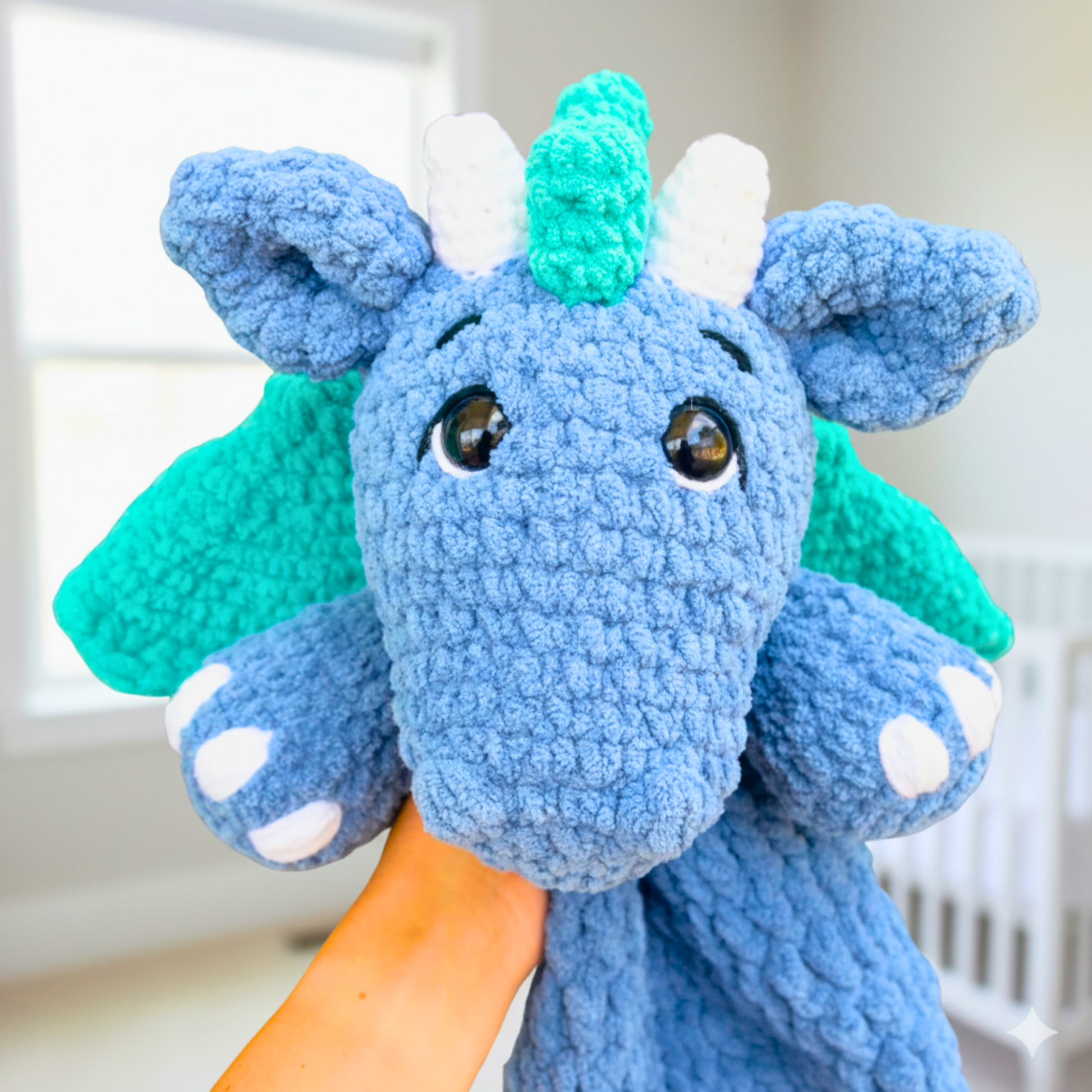 Hand holding a blue crocheted dragon plush toy with green wings and a horn in a bright room