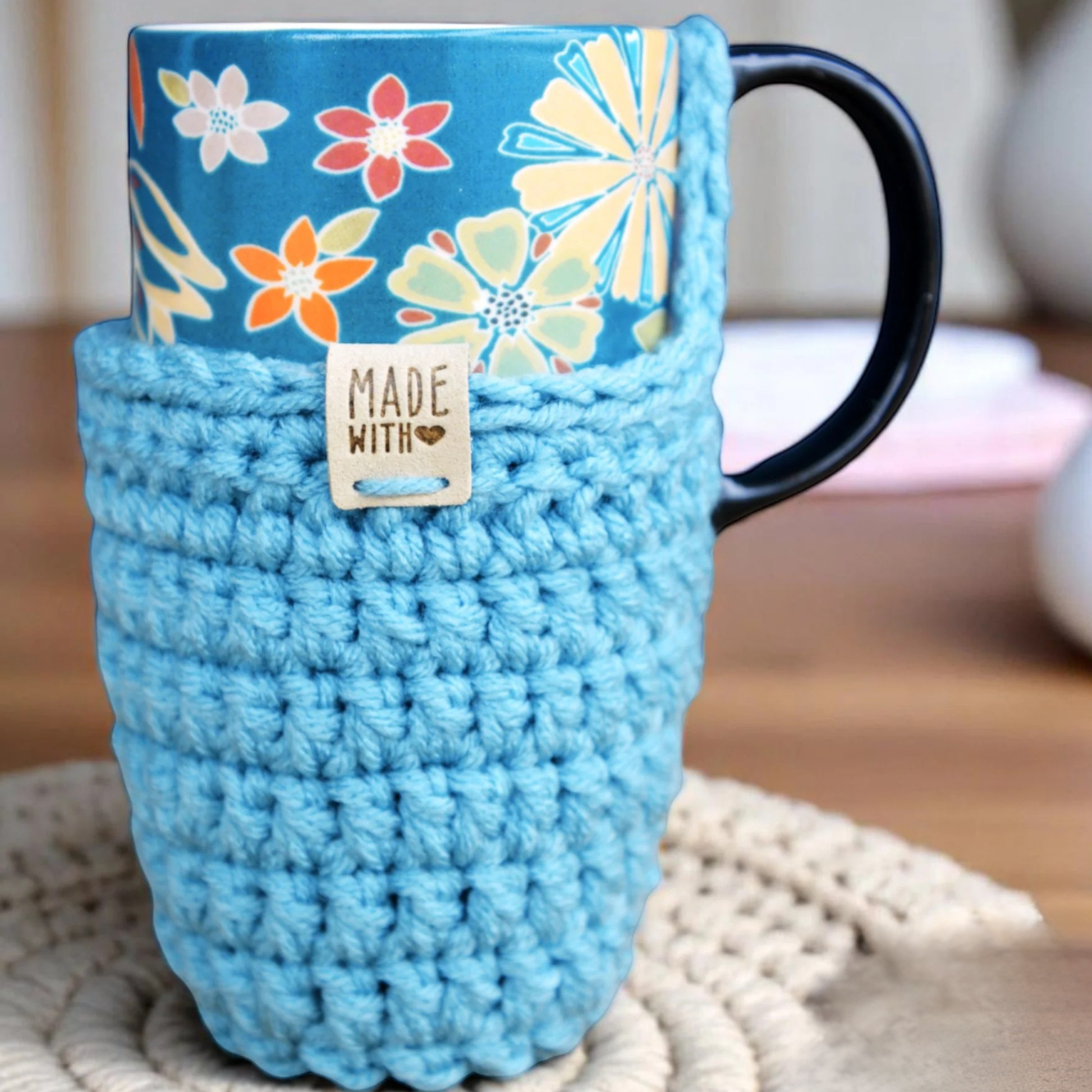 Colorful floral mug wrapped in handmade blue crochet cozy with 'Made With Love' tag