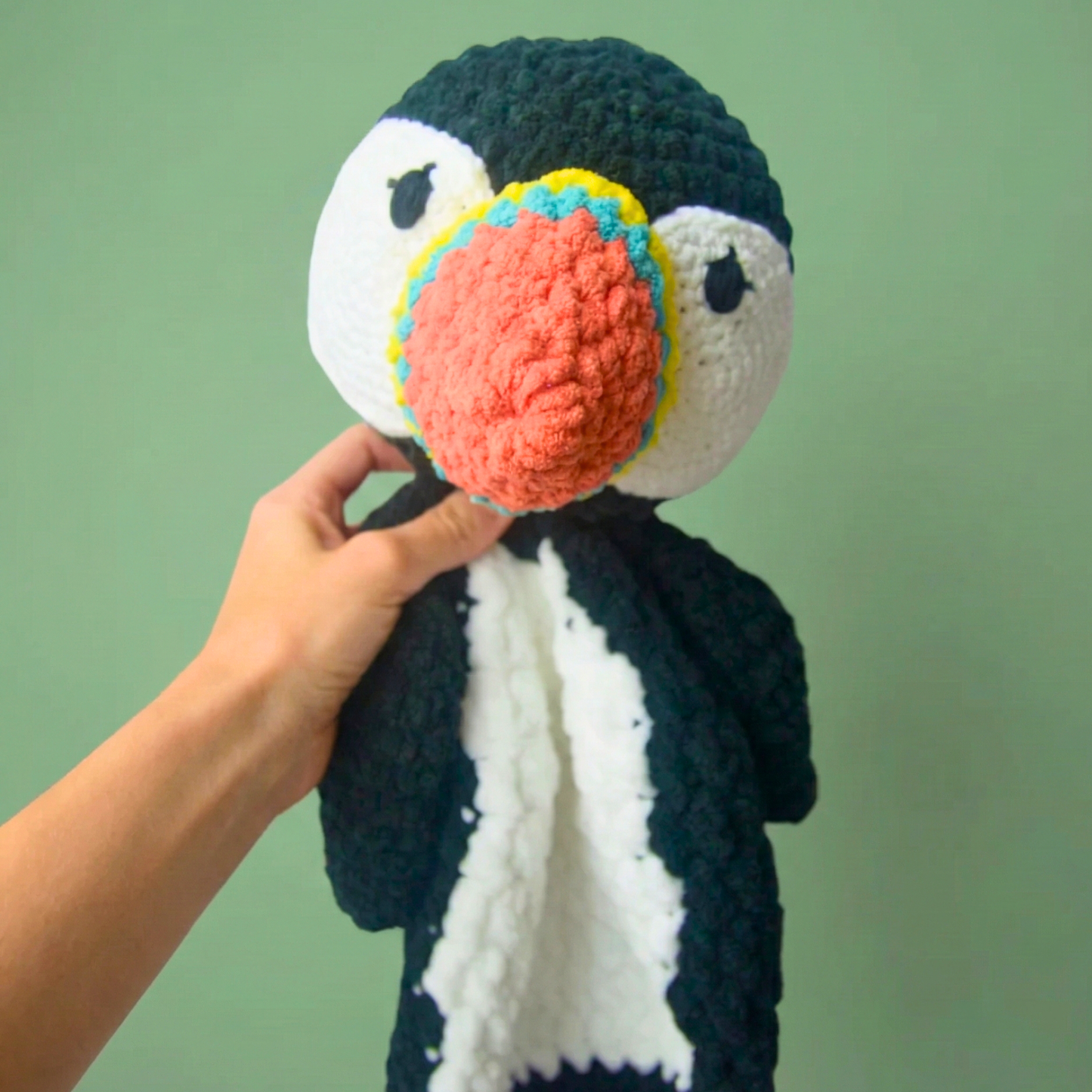 Hand holding a handmade plush penguin toy with black, white, and colorful beak against a green background