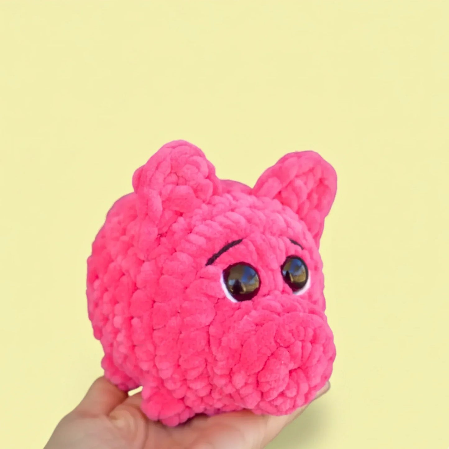 Hand holding a pink crochet pig plushie with big eyes on a yellow background