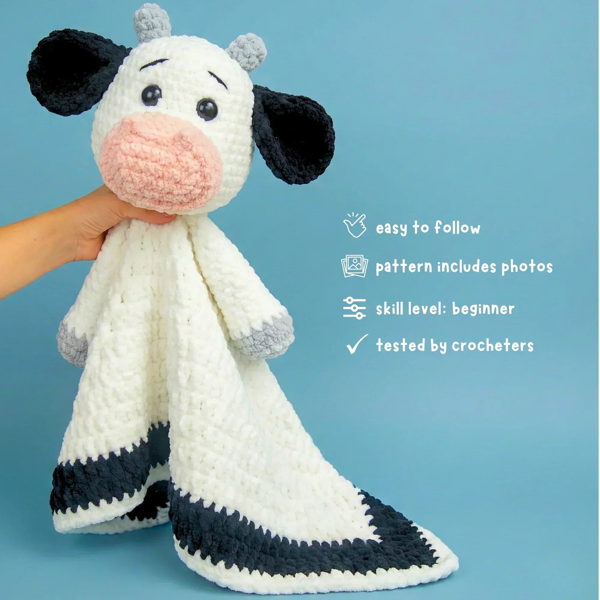 Hand holding crochet cow lovey plushie with black ears and white blanket on blue background