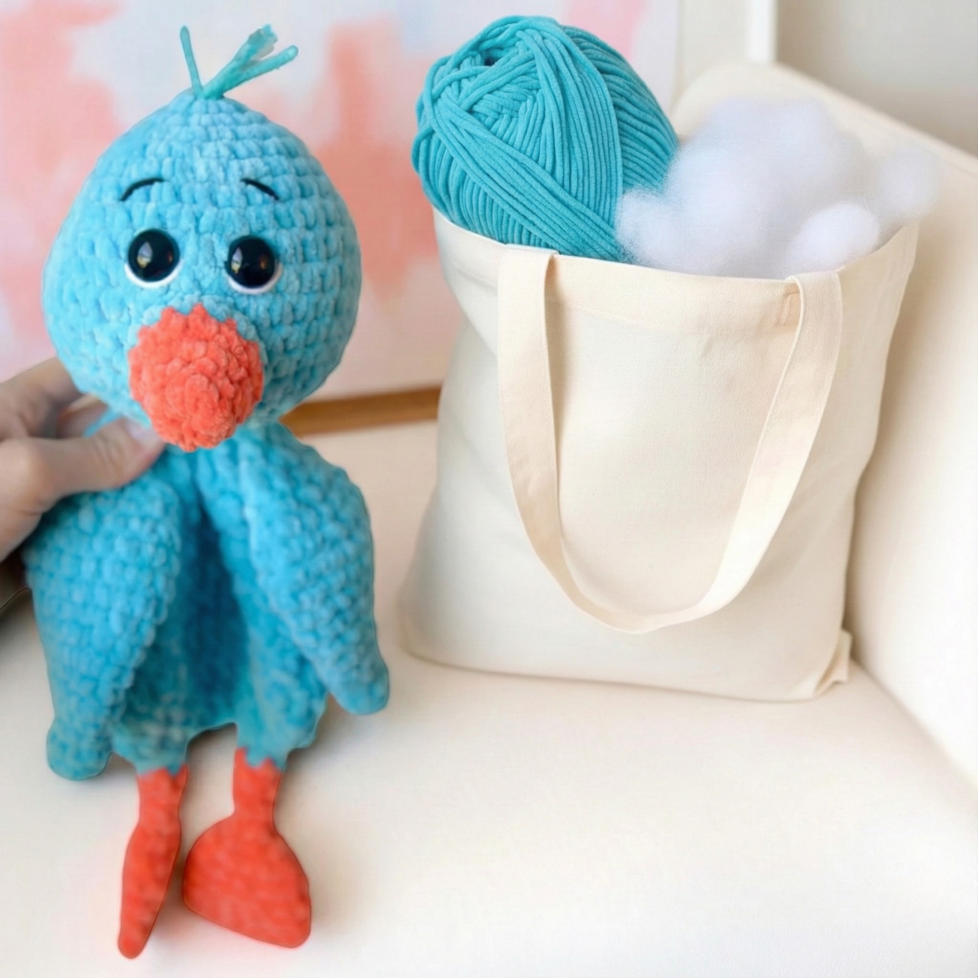 Hand holding a blue crocheted bird toy with orange beak and feet next to a tote bag with yarn and stuffing