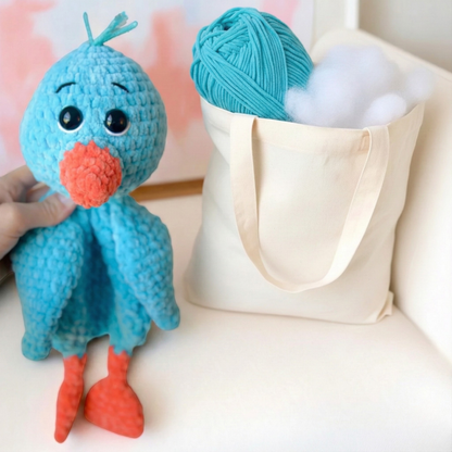 Hand holding a blue crocheted bird toy with orange beak and feet next to a tote bag with yarn and stuffing