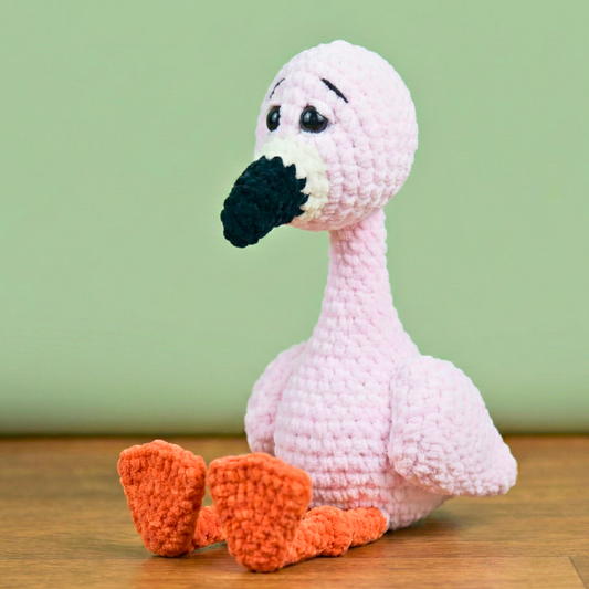 Handmade pink crocheted flamingo toy with black beak and orange feet on wooden surface