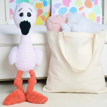 Pink crochet flamingo plushie with orange legs beside tote bag of yarn and stuffing