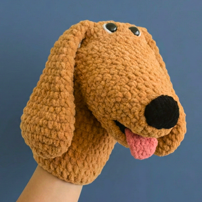 Hand puppet of a crocheted brown dog with floppy ears, black nose, and pink tongue on blue background