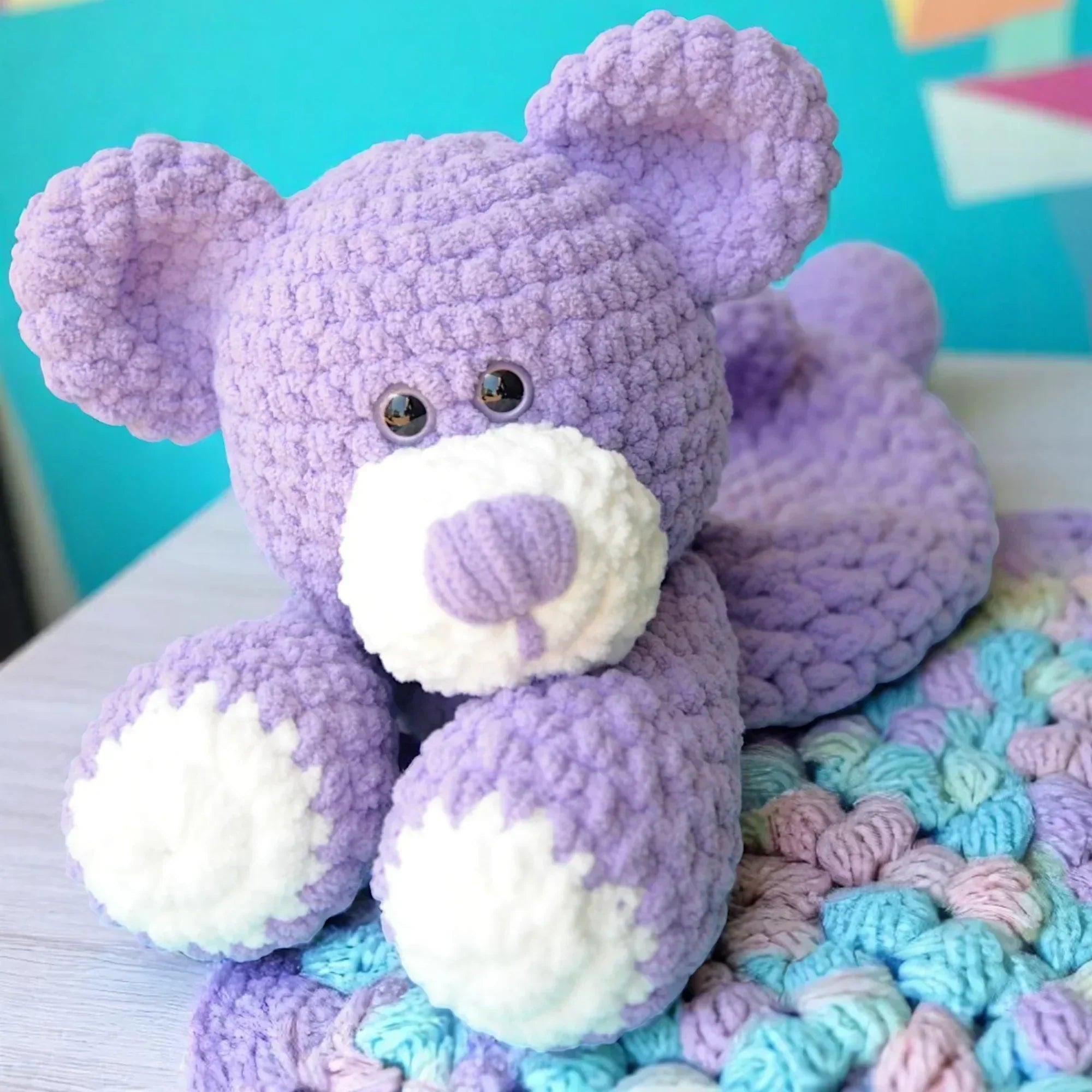 Purple crochet teddy bear plushie with white paws resting on a colorful crocheted blanket