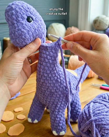 Hands crocheting a purple brontosaurus amigurumi plushie with yarn and crochet hook