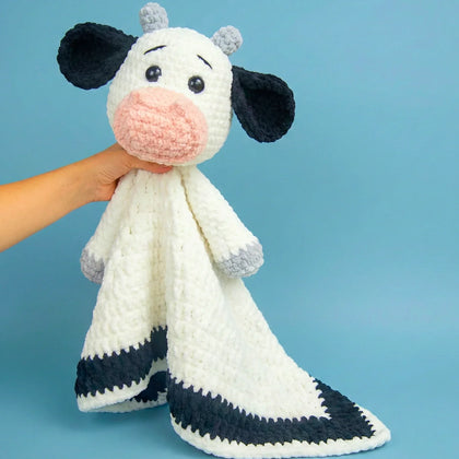 Hand holding crochet cow blanket with plush head, black ears, and white and black body on blue background.